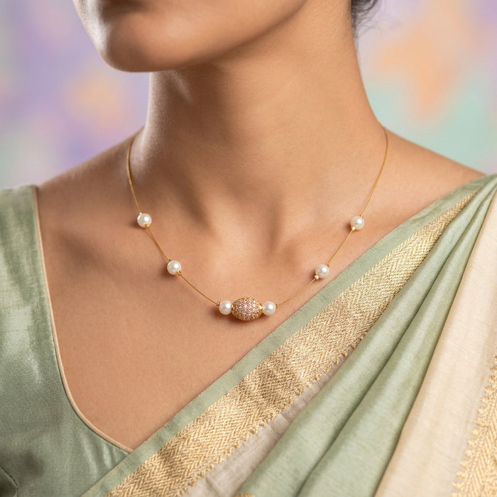 A delicate gold chain necklace displayed on a light gray fabric jewelry bust. The necklace features five white pearls spaced along the chain, with a central gold bead embellished with tiny clear stones.